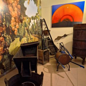 Strumenti in legno tradizionali per la produzione dell'aceto balsamico all'interno di una stanza del museo dell'aceto balsamico di Spilamberto, Modena, con un quadro di arte astratta blu e arancio appeso alla parete sul fondo
